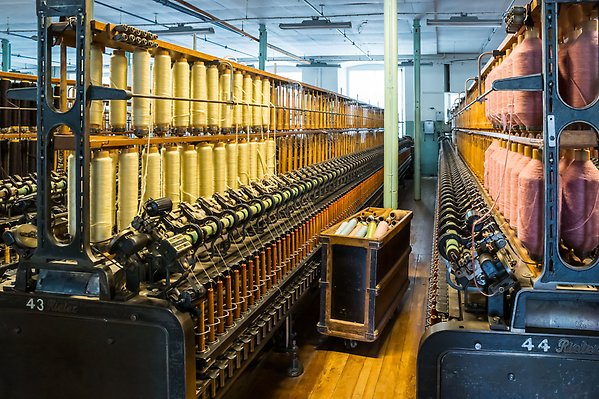 Gamla spinnerimaskiner står uppradade i en gammal fabrik. I maskinerna sitter gamla trådrullar.
